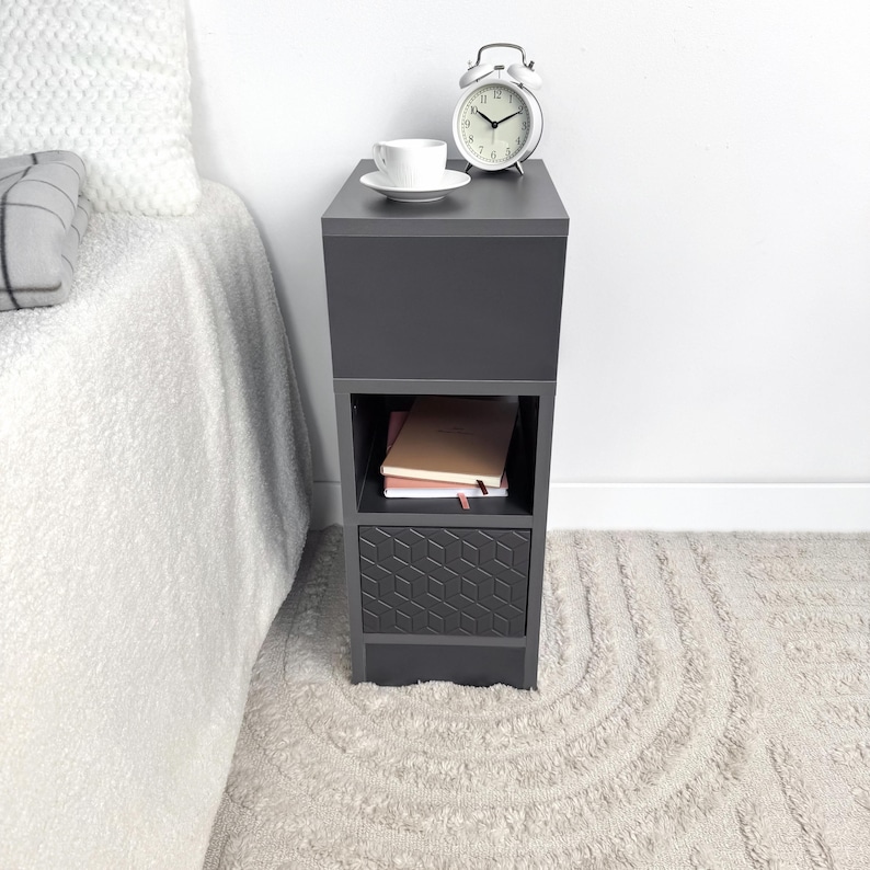 May include: A dark gray bedside table with a geometric patterned drawer. A white teacup and saucer sit on top, next to a white alarm clock. The table has an open shelf with books. The table is next to a bed with a white comforter.