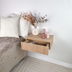May include: A light wood floating nightstand with a drawer that is open, revealing a pair of glasses. The nightstand is mounted on a white wall and has a vase with dried flowers and pink candles on top.