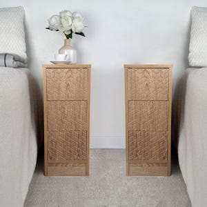 May include: Two matching wooden nightstands with geometric carved drawer fronts. Each nightstand is made of light-colored wood and has a top surface. A vase with white flowers and a cup and saucer sit on one nightstand.