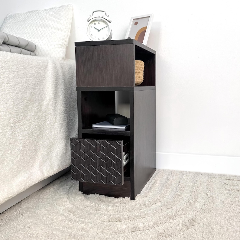May include: A dark brown nightstand with open shelves and a drawer with a geometric pattern. The nightstand has a clock, a framed picture, and a woven basket on top. A bed with a white comforter is next to the nightstand.