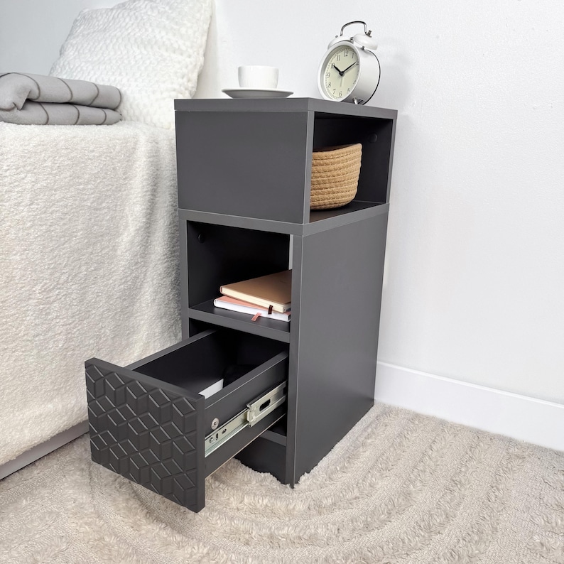 May include: A dark gray nightstand with an open drawer, a woven basket, and a white alarm clock. The drawer has a geometric pattern. A white teacup and saucer sit on top. The nightstand has two shelves and a drawer.