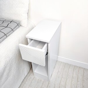 May include: A white bedside table with a drawer that has a spiral design. The drawer is open, revealing the interior. The table is next to a bed with a white comforter and a gray and white patterned blanket.