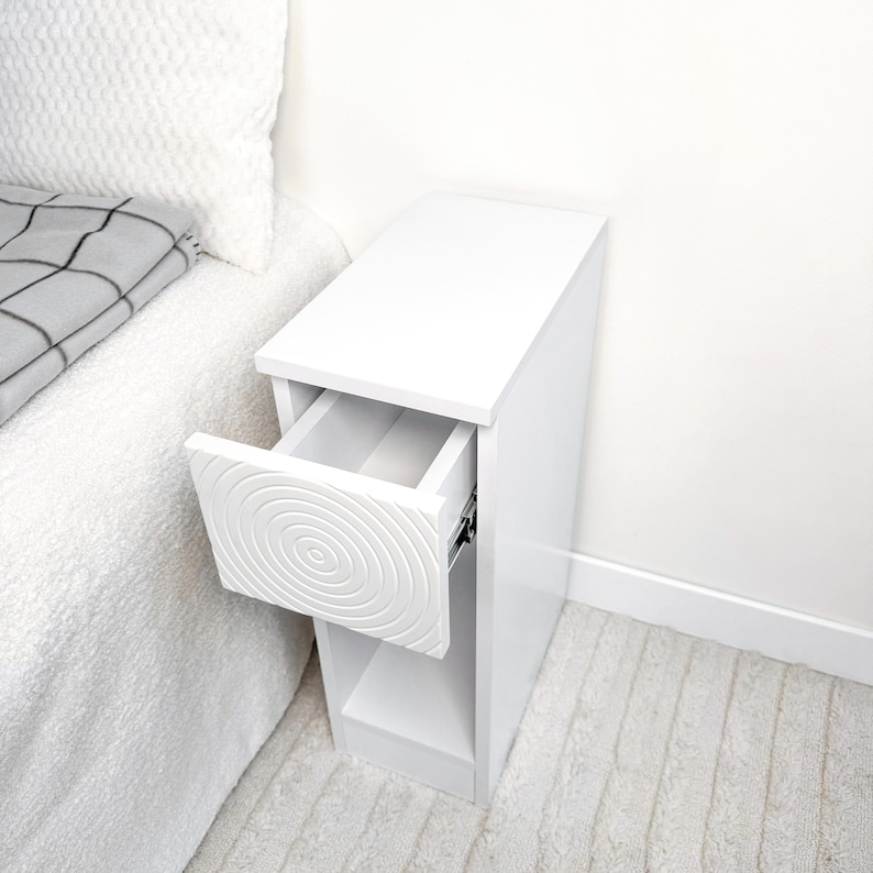 May include: A white bedside table with a drawer that has a spiral design. The drawer is open, revealing the interior. The table is next to a bed with a white comforter and a gray and white patterned blanket.