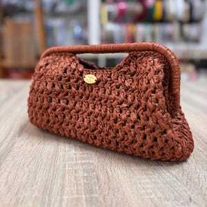 Puede incluir: Un bolso de mano tejido marrón con asa. El bolso tiene forma rectangular con bordes redondeados y una pequeña etiqueta dorada. La textura del bolso parece estar hecha de fibra natural.
