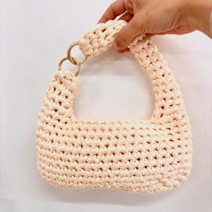 Può includere: Una borsa a mano color crema, realizzata a mano all'uncinetto, dalla forma strutturata. La borsa presenta una trama intrecciata, un manico arrotondato e dettagli con anelli metallici color oro. La borsa è tenuta da una mano su sfondo bianco.