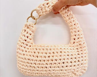 May include: A cream-colored, hand-crocheted handbag with a structured shape. The bag features a woven texture, a rounded handle, and gold-colored metal ring accents. The bag is held by a hand against a white background.