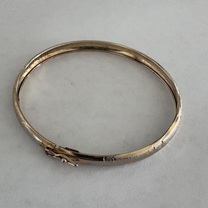 May include: A gold-tone metal hinged bangle bracelet with a textured, matte finish on the outer surface. The bracelet features a secure box clasp mechanism with a small side latch, creating a continuous oval shape when closed.