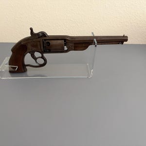 Puede incluir: Revólver antiguo con empuñadura de madera marrón oscuro y armazón metálico. El arma de fuego se exhibe sobre un soporte de acrílico transparente, mostrando su diseño histórico y artesanía. Se ven el cañón y el cilindro del arma.