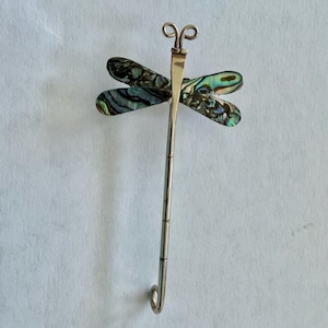 May include: A dragonfly-shaped brooch crafted from polished silver-toned metal. The four wings are inlaid with iridescent abalone shell featuring swirling patterns of green, blue, and black. The long, slender body has segmented detailing.