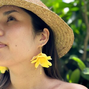 K&ouml;nnte beinhalten: Eine Person tr&auml;gt einen Strohhut und gelbe Blumenohrringe. Die Ohrringe sind aus Stoff und haben einen zarten, femininen Look.