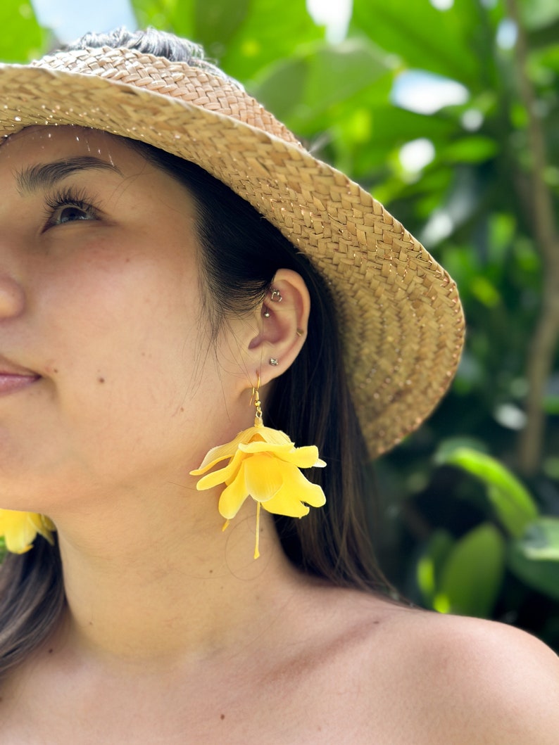 K&ouml;nnte beinhalten: Eine Person tr&auml;gt einen Strohhut und gelbe Blumenohrringe. Die Ohrringe sind aus Stoff und haben einen zarten, femininen Look.