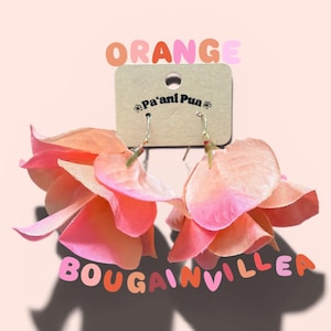 May include: A pair of orange and pink bougainvillea flower earrings. The earrings are made of layered fabric petals and hang from silver-colored hooks. The word "ORANGE" is above the earrings, and "BOUGAINVILLEA" is below.