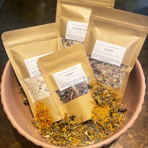 May include: A pink bowl filled with four small, brown paper bags of bath salts. Each bag has a white label with text. Dried flowers and herbs surround the bags. The labels read "Flower Chi" and "Purify".