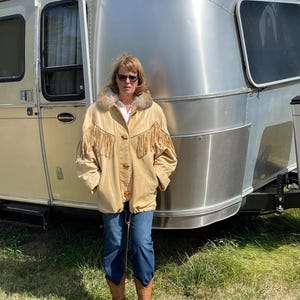 May include: A tan leather jacket with fringe detailing and a fur collar. The jacket has button closures and is worn with blue jeans and brown cowboy boots. An Airstream trailer is in the background.