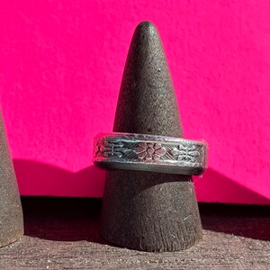 Puede incluir: Un anillo de plata con un intrincado diseño floral se encuentra sobre un soporte de anillo negro. El anillo se muestra sobre un fondo rosa brillante.