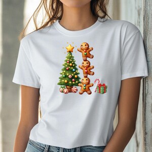 May include: White t-shirt featuring a festive Christmas design. The graphic includes a decorated Christmas tree, three gingerbread men stacked on top of each other, a candy cane, and a wrapped gift. The design is colorful and cheerful.