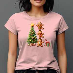May include: A light pink t-shirt featuring a Christmas design. The graphic includes a decorated Christmas tree, three gingerbread men stacked on top of each other, candy canes, and a wrapped gift.