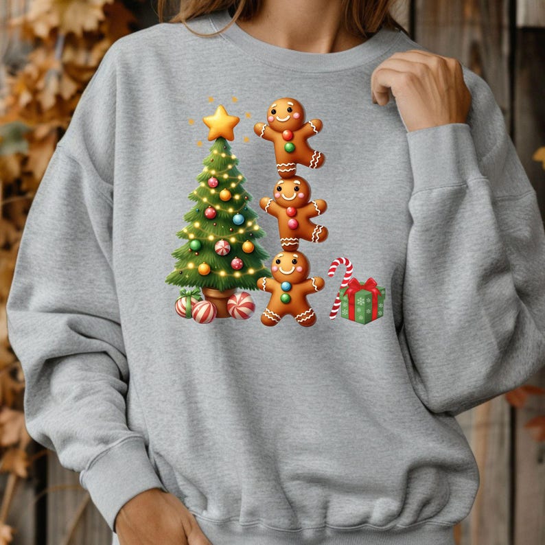 May include: A light gray sweatshirt featuring a Christmas design. The design includes a decorated Christmas tree, three gingerbread men stacked on top of each other, a candy cane, and a wrapped gift. The tree is adorned with lights and ornaments.