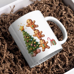 May include: White ceramic mug featuring a Christmas tree and gingerbread men design. The mug has a curved handle and is decorated with a star, candy canes, and a wrapped gift. The mug is placed in a white box with brown packing material.