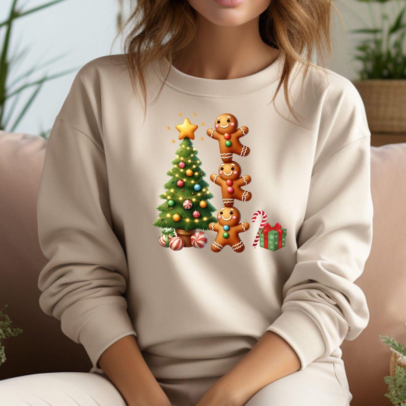 May include: Beige sweatshirt featuring a festive Christmas design. The graphic includes a decorated Christmas tree, a stack of gingerbread men, a wrapped gift, and a candy cane. The design is colorful and whimsical, perfect for the holiday season.