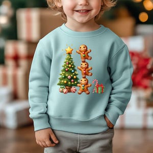May include: A light blue sweatshirt featuring a Christmas tree with ornaments, a star, and three gingerbread men. A candy cane and a wrapped gift are also visible. The sweatshirt is designed for children.