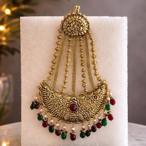 May include: Ornate gold-toned headpiece with intricate floral and geometric patterns. Features a central red gemstone and dangling red, green, and white beads. The headpiece is displayed on a white surface.
