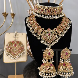 Puede incluir: Un conjunto de joyas de novia indias en tono dorado con piedras preciosas rosas y verdes. El conjunto incluye un collar, un choker, pendientes y un maang tikka.