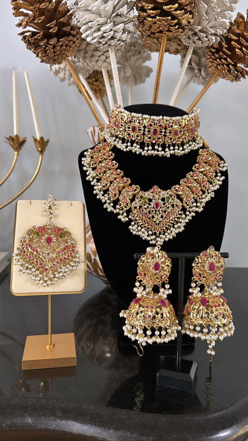 Puede incluir: Un conjunto de joyas de novia indias en tono dorado con piedras preciosas rosas y verdes. El conjunto incluye un collar, un choker, pendientes y un maang tikka.