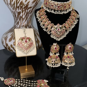 Puede incluir: Un conjunto de joyas de novia de oro con piedras rosas y verdes, perlas y una cadena delicada. El conjunto incluye un collar, pendientes, un maang tikka y un haar en capas.