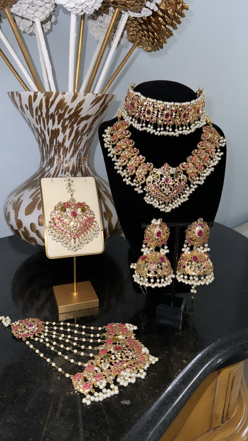 Puede incluir: Un conjunto de joyas de novia de oro con piedras rosas y verdes, perlas y una cadena delicada. El conjunto incluye un collar, pendientes, un maang tikka y un haar en capas.