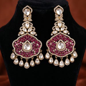 May include: A pair of ornate gold-tone chandelier earrings featuring deep pink oval-cut stones, clear teardrop-shaped stones, and small pavé-set crystals. The earrings have a central teardrop stone surrounded by pink stones and dangling accents.