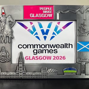 May include: A rectangular metal picture frame featuring embossed Glasgow landmarks, including the Duke of Wellington statue with a traffic cone, a ship, and the Scottish flag. The top includes a pink plaque reading PEOPLE MAKE GLASGOW.