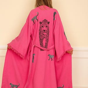 Puede incluir: Una bata rosa vibrante con un diseño de estampado de leopardo. La bata presenta una gran cara de leopardo en la espalda y figuras de leopardo más pequeñas dispersas por la tela. Tiene bordes con flecos y un cinturón a juego.