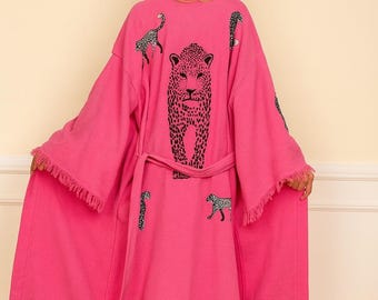 May include: A bright pink long-sleeved robe featuring a large black leopard print on the back and smaller leopard motifs scattered across the fabric. The garment includes a matching tie belt at the waist and frayed edges along the hem.