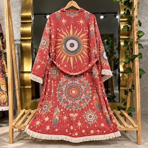 May include: A long-sleeved, knee-length red robe featuring a prominent sun graphic on the back, a large circular mandala pattern on the lower section, and white fringe trim along the hem and sleeve cuffs, hanging on a wooden hanger.