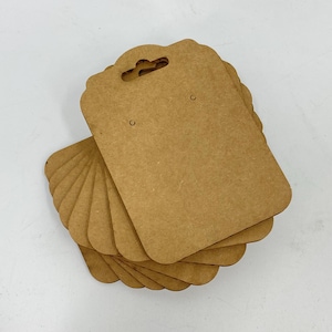 Puede incluir: Una pila de tarjetas de exhibición de pendientes marrones con bordes festoneados. Cada tarjeta tiene un pequeño agujero para pendientes y un agujero para colgar. Las tarjetas están hechas de un material de papel grueso y texturizado.