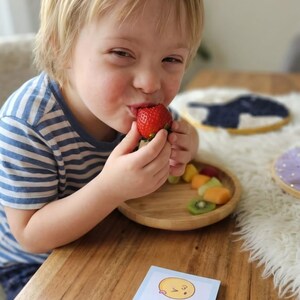 Puede incluir: Un conjunto de tarjetas de actividades educativas con ilustraciones coloridas y texto que incluye KISS KISS y COLOR SORT, junto a un pequeño plato de madera que contiene trozos de fruta fresca como fresas, kiwi y melón.
