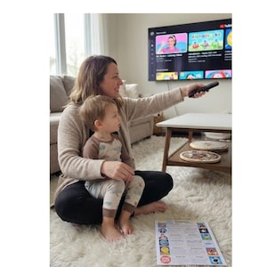 May include: A black television remote control held in a hand, pointed towards a wall-mounted television screen displaying a YouTube interface with colourful educational video thumbnails, including one labelled Alphablocks with letter characters.