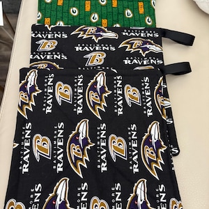 May include: A set of square fabric pot holders featuring two distinct designs. The top two are green with a repeating Green Bay Packers logo and text. The bottom two are black with a repeating Baltimore Ravens logo and text.