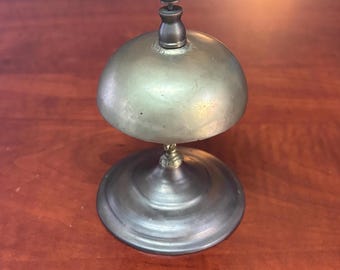May include: A vintage brass bell with a dome-shaped top and a round base. The bell has a small handle on top and a clapper inside. The bell is likely used for calling attention or signaling.