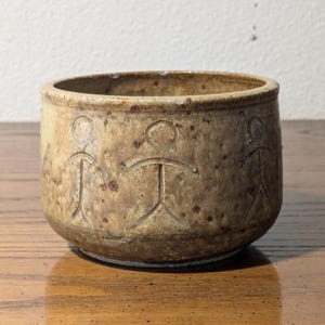 May include: A small, round, ceramic bowl with a speckled, tan exterior. The bowl features three etched figures around the exterior. The figures have a simple stick-figure design. The bowl sits on a wooden surface.