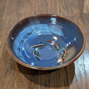 May include: A ceramic bowl with a blue interior featuring painted fish. The bowl has a brown rim and exterior. The fish are painted in shades of red, green, and white, creating a decorative aquatic theme.