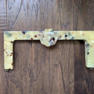 May include: A rectangular purse frame in a speckled cream and brown pattern. The frame has a decorative center piece and small metal rivets at the base of each handle. The frame is set against a wooden background.