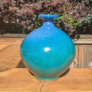 May include: A handcrafted ceramic vase with a rounded body and a narrow neck. The vase features a gradient of blue and teal hues, with a speckled texture. The vase is displayed on a wooden surface, showcasing its unique design and craftsmanship.