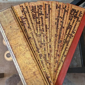 May include: An antique, open book with gold-colored pages and black text. The pages are fanned out, revealing a red spine. The book's cover is gold with ornate patterns.