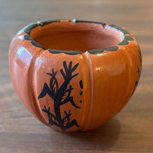 May include: A small, orange ceramic pumpkin-shaped bowl with a black rim and black decorative accents. The bowl has a carved pumpkin design and is likely used for Halloween or fall decor.