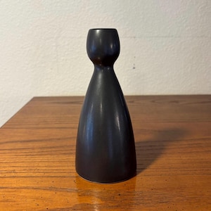 May include: A matte black candle holder with a smooth, tapered silhouette. The object features a wide, circular base that narrows into a slender neck before expanding slightly at the top to form a cup-shaped opening for a single taper candle.
