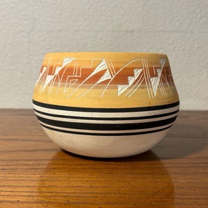 May include: A handcrafted ceramic bowl with a rounded shape. The bowl features a cream-colored base, with black and white stripes. The upper portion is yellow with a geometric design in white and orange. A decorative piece.