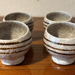 May include: Four small, round, ceramic bowls with a speckled gray finish. Each bowl has three horizontal bands of dark brown around the middle. The bowls sit on a wooden surface.