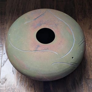 May include: A round, ceramic decorative bowl with a central hole. The bowl has a mottled green and reddish-brown finish with etched, curved lines. The design suggests a stylized fish or aquatic creature. The bowl sits on a wooden surface.